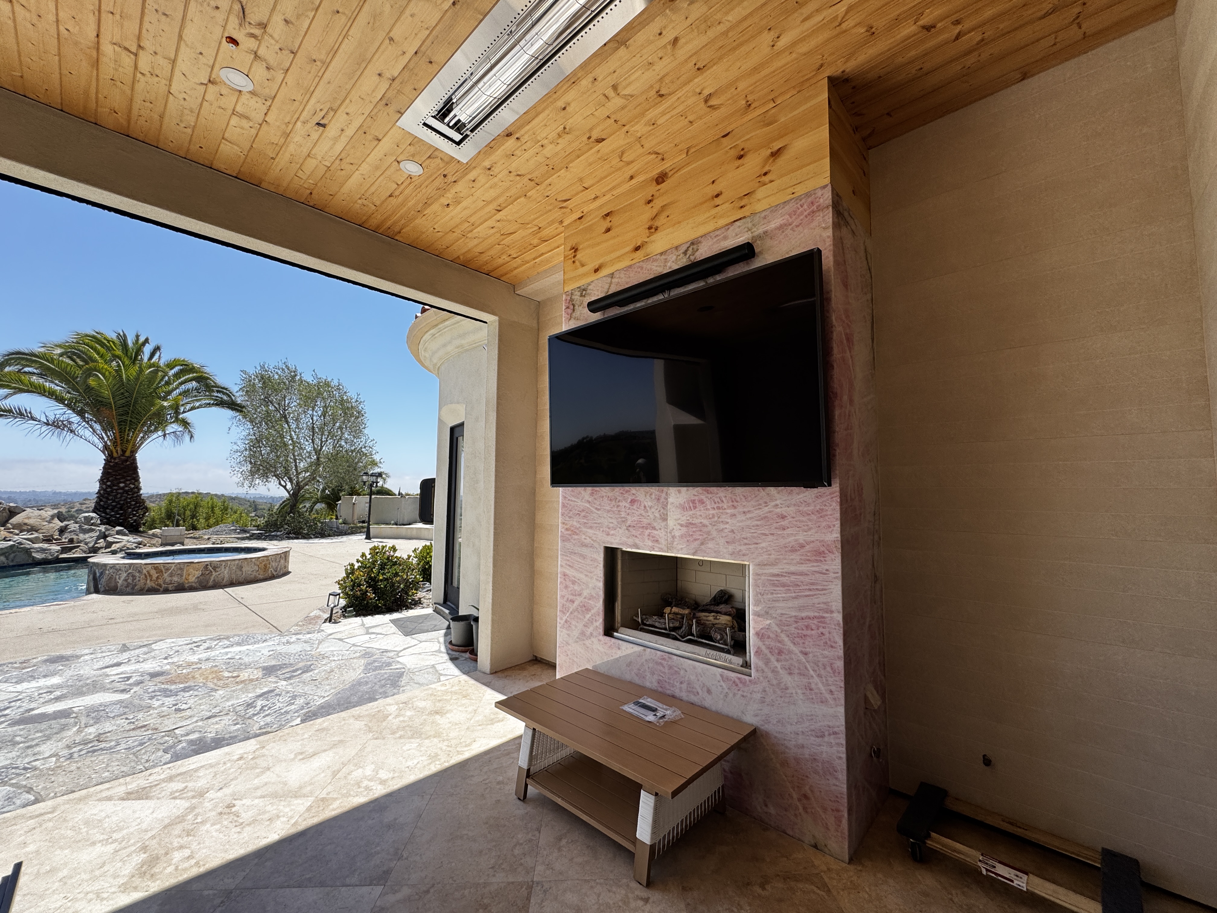 MKR Systems completed outdoor patio TV installation — large TV on pink marble wall above Heatilator fireplace, pool view, San Diego