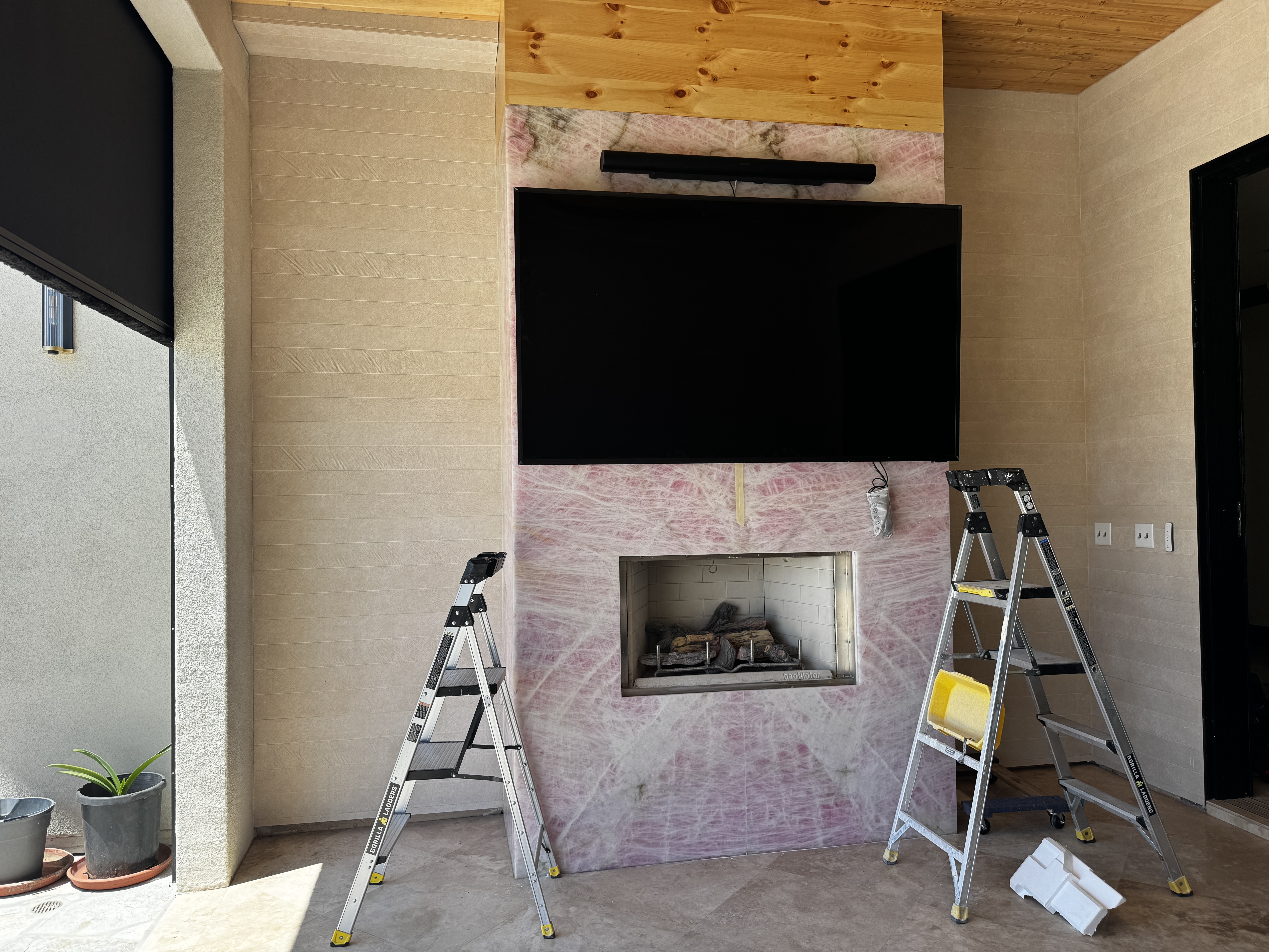 MKR Systems outdoor patio TV installation in progress — pink marble fireplace wall, two Gorilla Ladders, large format TV mounted